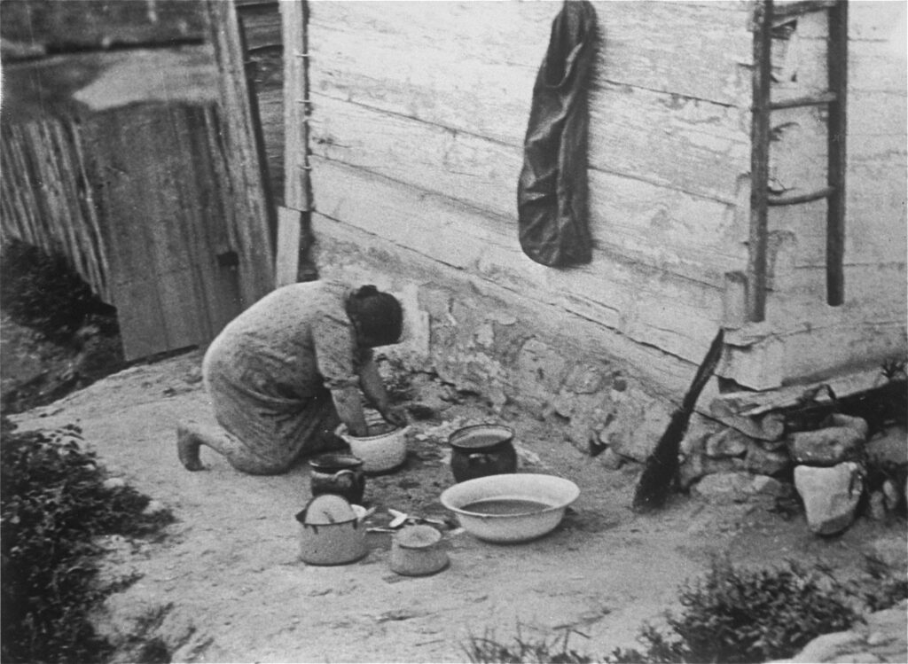 In the town of Kazimierz Dolny before the Nazi invasion, an unidentified Jewish woman kneels outside next to a wooden building, washing items in a basin. Several pots and a bowl are nearby. A jacket hangs on the wall beside a simple wooden ladder.