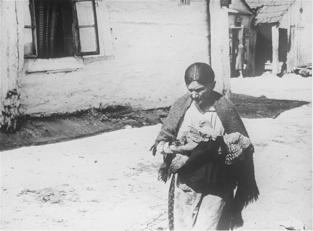 An unnamed Jewish woman is walking outside in Kazimierz Dolny. In her arms, she holds a chicken partially wrapped in a cloth. She is wearing a shawl, and behind her are old buildings with wooden roofs and open windows.