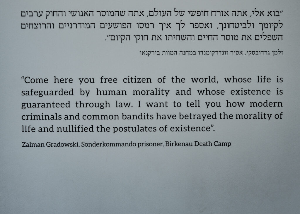 Print on a wall in Hebrew and English. The English text reads: ""Come here you free citizen of the world, whose life is safeguarded by human morality and whose existence is guaranteed through law. I want to tell you how modern criminals and common bandits have betrayed the morality of life and nullified the postulates of existence". Zalman Gradowski, Sonderkommando prisoner, Birkenau Death Camp"