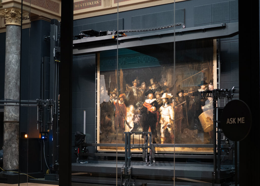 An image of the painting The Night Watch by Rembrandt undergoing restoration work behind glass at the Rijksmuseum