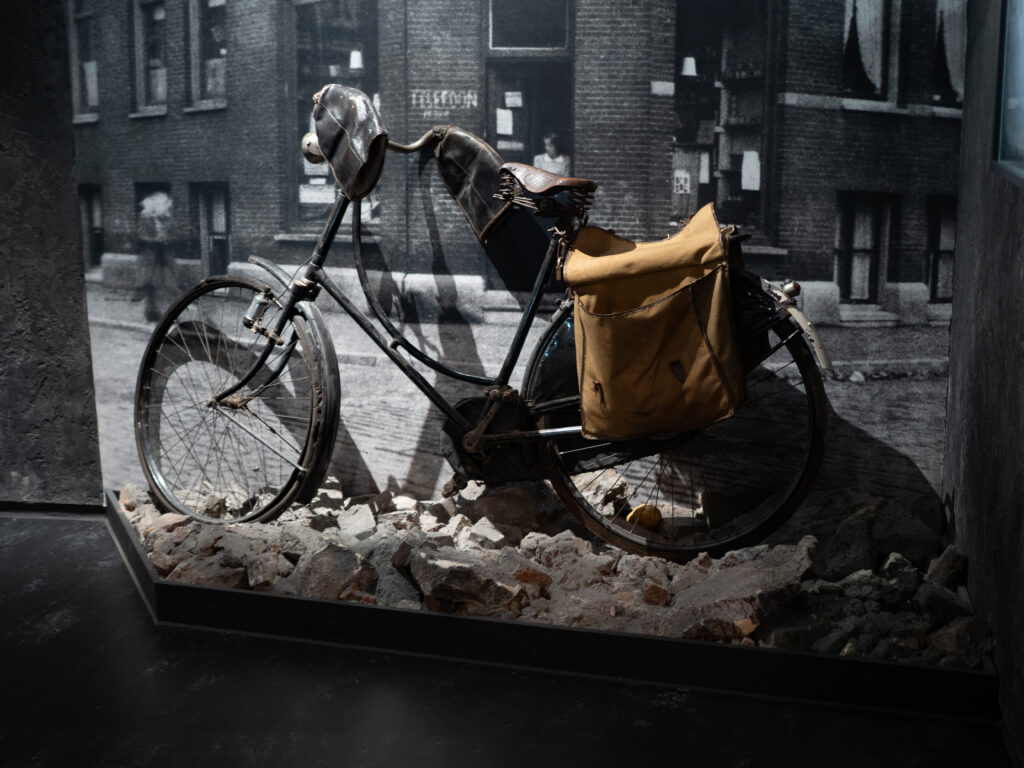 A bike belonging to a member of the Netherland resistance during WWII, on exhibit at the Resistance Museum in Amsterdam. The big is black with a brown letter seat, and a sack is attached at the back. It is displayed against a blown of image of Amsterdam during the war in black and white.