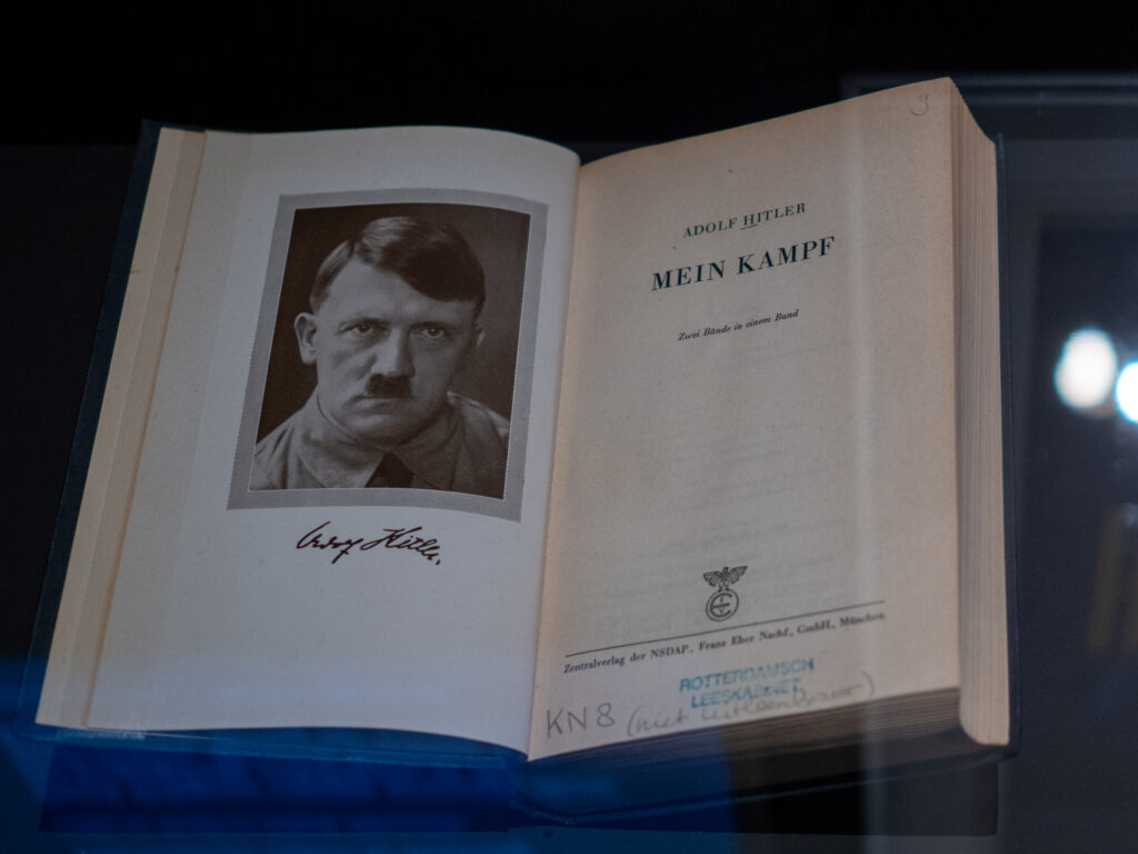A copy of Mein Kampf is open to the title page, with a photo of a menacing looking Hitler on the left, and the title on the right.