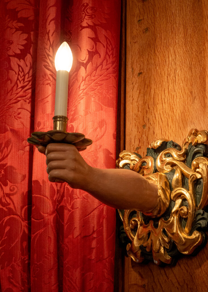 This wall sconce has gold filigree on the wall portion, and a fake arm extends out to hold a candle (electric now). There is a red damask curtain behind the candle, and the wall the sconce is attached to is wood.