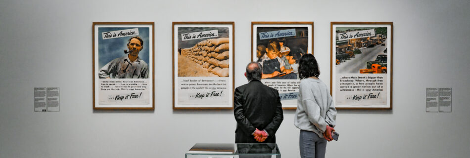 An image of a man and a woman standing before the Selling Point part of the Rijksmuseum's exhibit "American Photography". There is a green sign that says Selling Point in the top left corner. There are 4 colourful posters on the wall, each selling a particular aspect of America. They all have the header "This is America" with the tagline "Keep it Free".
